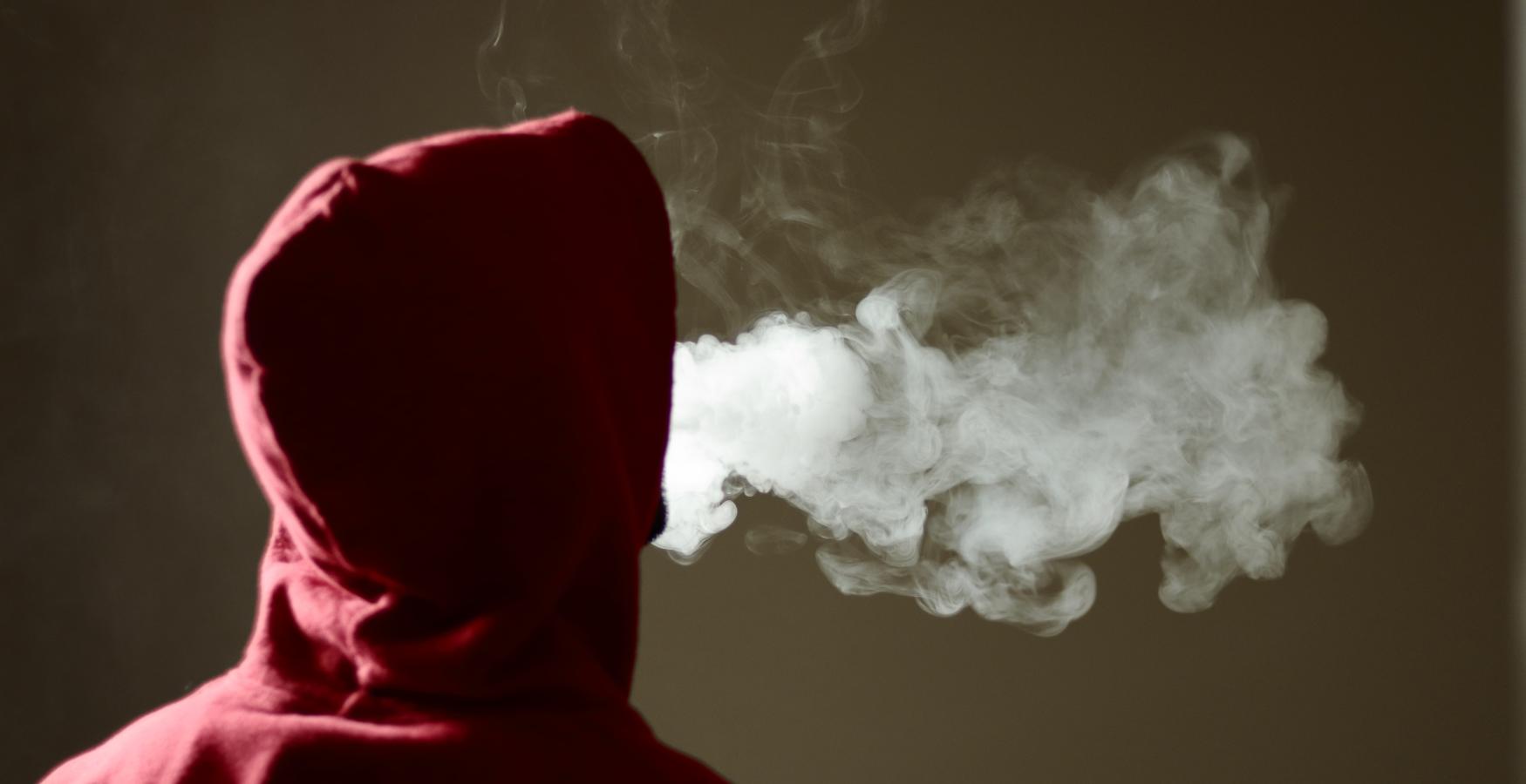 The back of the head and shoulders of a person wearing a hoodie, with a cloud of exhaled smoke.